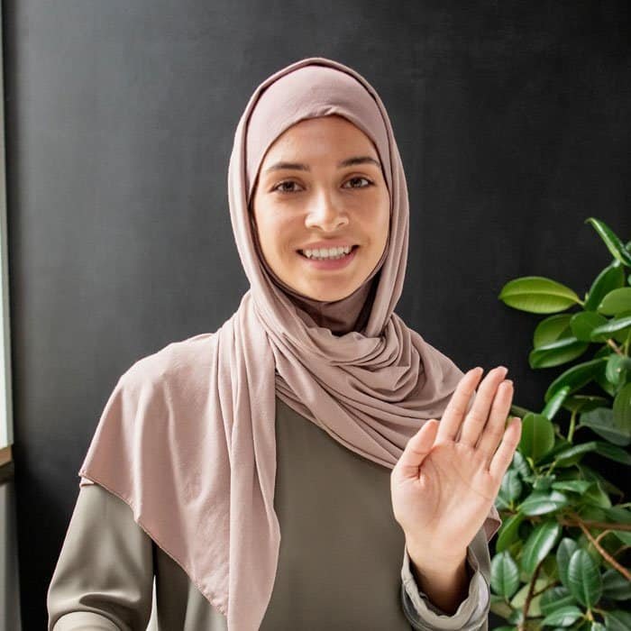 Woman in hijab waving indoors with green plant in background.